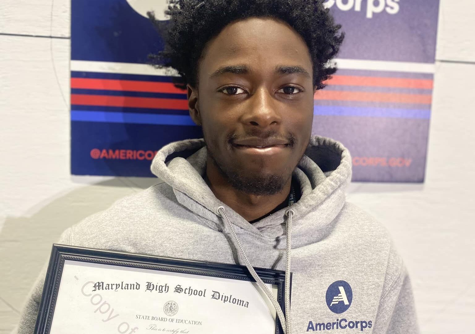 young man wearing an AmeriCorp sweatshirt holding high school diploma