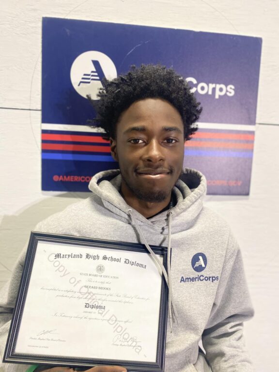 young man wearing an AmeriCorp sweatshirt holding high school diploma