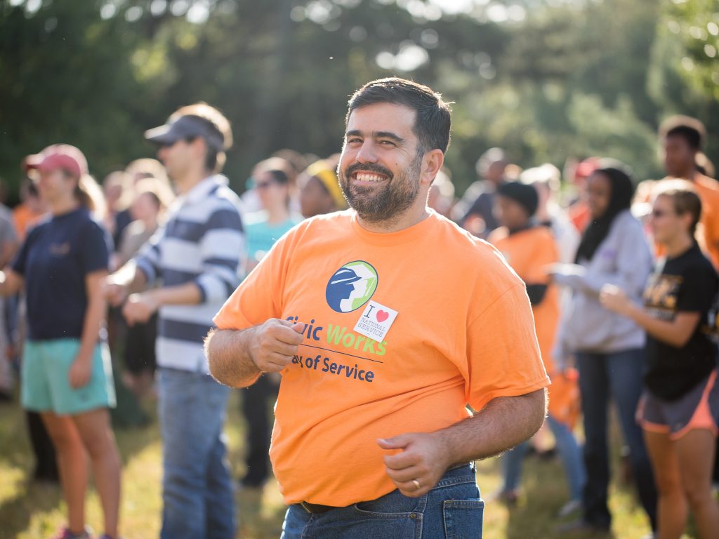 volunteer in an orange civic work shirt smiles while standing outside with a group of volunteers stands behind them