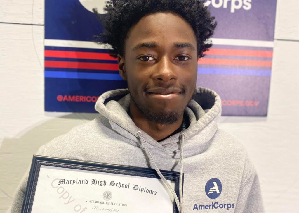 young man wearing an AmeriCorp sweatshirt holding high school diploma